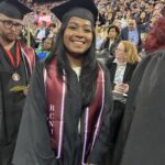 A smiling graduate in a black cap and gown with a maroon stole labeled RCNJ stands among classmates at a crowded graduation ceremony. An audience watches from the stands behind her.