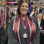 A smiling graduate in a cap and gown stands at a commencement ceremony. She wears maroon stoles, a medallion labeled EOF, and a decorated graduation cap, with an audience and other graduates in the background.