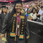 A smiling graduate wearing a cap and gown with a Ramapo College stole stands indoors at a commencement ceremony, with a crowd of people and other graduates in the background.