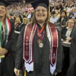 A smiling graduate wearing a cap and gown stands at a ceremony, adorned with maroon and white stoles, medals, and honors. The background shows a crowded auditorium with seated spectators.