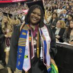 A smiling graduate in cap and gown stands indoors at a Ramapo College ceremony, wearing honor cords, stoles, and holding a Jamaican flag, with a crowd of seated people in the background.