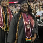 Two women wearing graduation caps, gowns, and honor cords smile at a graduation ceremony. The background shows an audience seated in an arena and a banner reading School of Social Work.