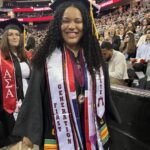 A smiling graduate in a cap and gown stands at a ceremony, wearing stoles that read First Generation and EOF. People in the audience and other graduates are visible in the background.