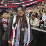 A graduate in cap and gown, adorned with stoles, medals, and honor cords, smiles at a commencement ceremony. An audience fills the arena behind, with “Ramapo College of New Jersey” displayed on a screen above.