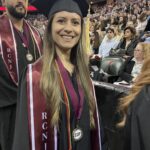 A smiling graduate in a cap and gown stands indoors at a commencement ceremony, wearing RCNJ and EOF medallions and a tassel. Other graduates and seated audience members are visible in the background.