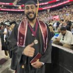 A smiling graduate in a black cap and gown with maroon stole and medallion gives a thumbs up at an indoor graduation ceremony, holding a diploma. The audience and other graduates are visible in the background.