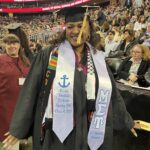 A smiling graduate in a cap and gown stands at a ceremony. She wears honor stoles with embroidery and a kente cloth. People are seated in the background under signs for “School of Social Science and Human Services.”.