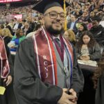 A graduate in cap and gown stands and smiles at a commencement ceremony. He wears multiple honor cords and a sash with RCNJ. Behind him, an audience of family and friends watches from the stands.
