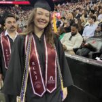 A smiling graduate in a cap and gown stands indoors at a commencement ceremony, wearing multiple honor cords and a Ramapo College of New Jersey stole. Rows of seated graduates and audience members fill the background.