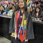 A young woman in a graduation cap and gown stands smiling, wearing multiple honor cords and stoles. Behind her, people sit in rows of seats at a crowded indoor graduation ceremony.