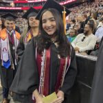 A smiling graduate in a cap and gown stands indoors at a commencement ceremony, wearing honor cords and a maroon stole, with other graduates and an audience seated in the background.