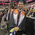 A smiling graduate in a cap and gown stands indoors at a commencement ceremony, holding a small flag. His stole reads NURSING, and he is surrounded by other graduates and an audience in the background.