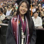 A young woman wearing a graduation cap and gown smiles at the camera. She has medals and cords around her neck, and there is a crowd of people seated in the background at a graduation ceremony.