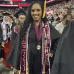A graduate in a black cap and gown, decorated with multiple honor cords and a maroon stole, smiles at a commencement ceremony. Crowds fill the arena, and Ramapo College of New Jersey is visible in the background.