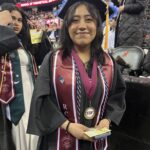 A graduate in a cap and gown smiles at a graduation ceremony, wearing maroon and white stoles, a medal, and holding diplomas. Other graduates and audience members are visible in the background.