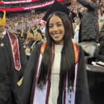 A young woman in a graduation cap and gown is smiling at a graduation ceremony. She wears a sash labeled Nursing and Ramapo College of New Jersey with a crowd and banners visible in the background.
