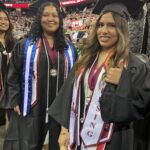 Two graduates in caps and gowns smile at a commencement ceremony. Both wear multiple honor cords and stoles, one with a Nursing sash, giving a thumbs up. A crowd and stage are visible in the background.