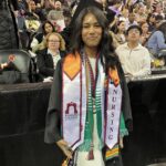 A smiling graduate in a cap and gown stands in front of a large seated crowd. She wears colorful stoles, including one with Nursing and Ramapo College on it, and has multiple honor cords around her neck.