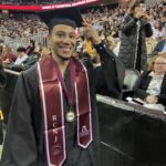 A graduate in a black cap and gown smiles while holding his tassel at a ceremony. He wears a maroon stole with RCNJ and EOF on it. People are seated behind him in the audience at an indoor arena.
