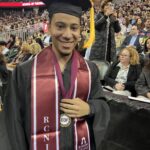 A smiling graduate in a cap and gown with an EOF medallion and RCNJ stole stands in an arena, with an audience seated behind him and red banners reading Ramapo College of New Jersey visible.