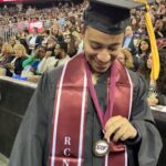 A student in graduation attire smiles and looks down at their EOF medallion during a ceremony. They wear a maroon stole with “RCNJ” and “Ramapo College” printed, with an audience seated in the background.
