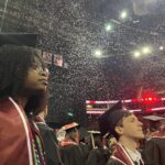 Graduates in caps and gowns stand together indoors as confetti falls from above. A woman in the foreground looks upward thoughtfully, and a man nearby looks ahead. Stadium lights and congratulatory signs are visible in the background.