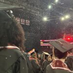 Graduates in caps and gowns stand together at a commencement ceremony indoors, with confetti falling from above and banners hanging in the background. The atmosphere is festive and celebratory.