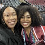 Two women smile and pose for a selfie at a graduation ceremony. One wears a black graduation gown, and the other wears a cap, gown, honor cords, and holds a diploma. Rows of empty chairs are in the background.
