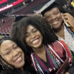 Three women in graduation gowns and caps smile for a selfie inside an arena. One holds a diploma and wears multiple honor cords; another is talking on a phone. The background shows rows of empty red seats.
