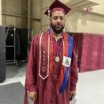 A graduate in a maroon cap and gown stands indoors, wearing a medal, colorful sash, and cords. The gown has RCNJ on a stole. The background includes red padding and beige walls.