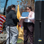 A person in a black hoodie receives a certificate from a woman in sunglasses and a pink vest, standing on an outdoor stage with American and New Jersey flags in the background.