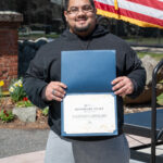 A man with glasses and a goatee smiles while holding an Honorary Staff certificate outdoors. An American flag and spring flowers are visible in the background.