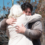 An older adult with white hair and a light coat hugs a younger person outdoors. The younger persons eyes are closed and they smile, both arms wrapped around the older adult. Leafy branches are visible in the background.