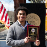 A young man smiles and holds a plaque standing outdoors in front of a podium with Ramapo College insignia and two American flags visible in the background.