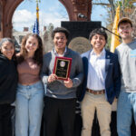 Five young adults stand together outdoors, smiling at the camera. The person in the center holds a plaque. There are flags, a podium, and a college emblem in the background, suggesting an award ceremony.