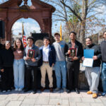 A group of eight young adults stand outdoors, smiling and posing together. Two hold plaques, and one holds a certificate. There is a stone arch and trees in the background, with flags and clear, sunny weather.