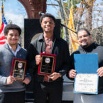 Three men stand outdoors, smiling and holding awards. Two hold plaques with EDP logos, and the third holds an Honorary Staff certificate. U.S. flags and trees are in the background on a sunny day.