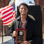 A young man smiles while holding a plaque for the EOF Emerging Leader Award. He stands outdoors in front of a U.S. flag and a decorative stone arch, dressed in a black jacket and patterned shirt.