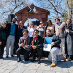 A group of people pose outdoors, smiling in front of a brick arch. Two people kneel in front holding award plaques, while others stand behind them, some holding certificates. It’s a sunny day with trees in the background.