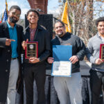 Four men stand outdoors, smiling and holding awards. Two men hold plaques, one holds a certificate, and another points at a plaque. Flags and trees are visible in the background on a sunny day.
