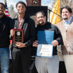 Four men stand outdoors, smiling at the camera. Two of them hold awards—one holds a plaque and the other a certificate. There is a podium and flags in the background.
