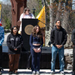 A group of seven young people stand in a row outdoors in front of a podium, where a woman is speaking. The scene appears to be part of an organized event or ceremony on a sunny day.