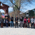 A diverse group of people stand in a line outdoors near a brick arch, with an American flag and speaker setup behind them. Trees and a modern building are visible in the background on a sunny day.