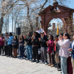 A group of people stands in a line outdoors in front of a brick archway, clapping and facing the left. Trees with bare branches are in the background, and its a sunny day.