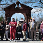 A diverse group of people stands outdoors in front of a decorative stone arch, listening attentively. Trees with sparse leaves and a blue sky are visible in the background.