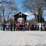 A large group of people stands outdoors in front of a decorative arch, clapping and facing the camera on a sunny day, with trees and a modern building in the background.