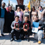 A group of thirteen people pose outdoors, some standing and some kneeling in front. Two people kneeling hold award plaques, while another holds a certificate. The group is smiling and dressed in business casual attire. Flags are visible behind them.