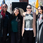 Four women stand side by side smiling outdoors in front of a podium with a college seal, with American and yellow flags in the background, dressed in coats and sunglasses on a sunny day.
