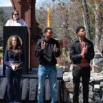 Five people stand outdoors on a stage, some clapping, with a woman speaking at a podium behind them. An American flag and trees are visible in the background on a sunny day.