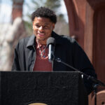 A person with short curly hair speaks into a microphone at an outdoor podium on a sunny day, wearing a black jacket over a red collared shirt. Trees and a building are visible in the background.